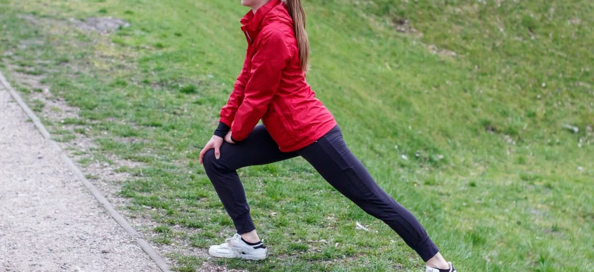 Woman in red jacket stretching in a park, illustrating outdoor fitness and healthy lifestyle.